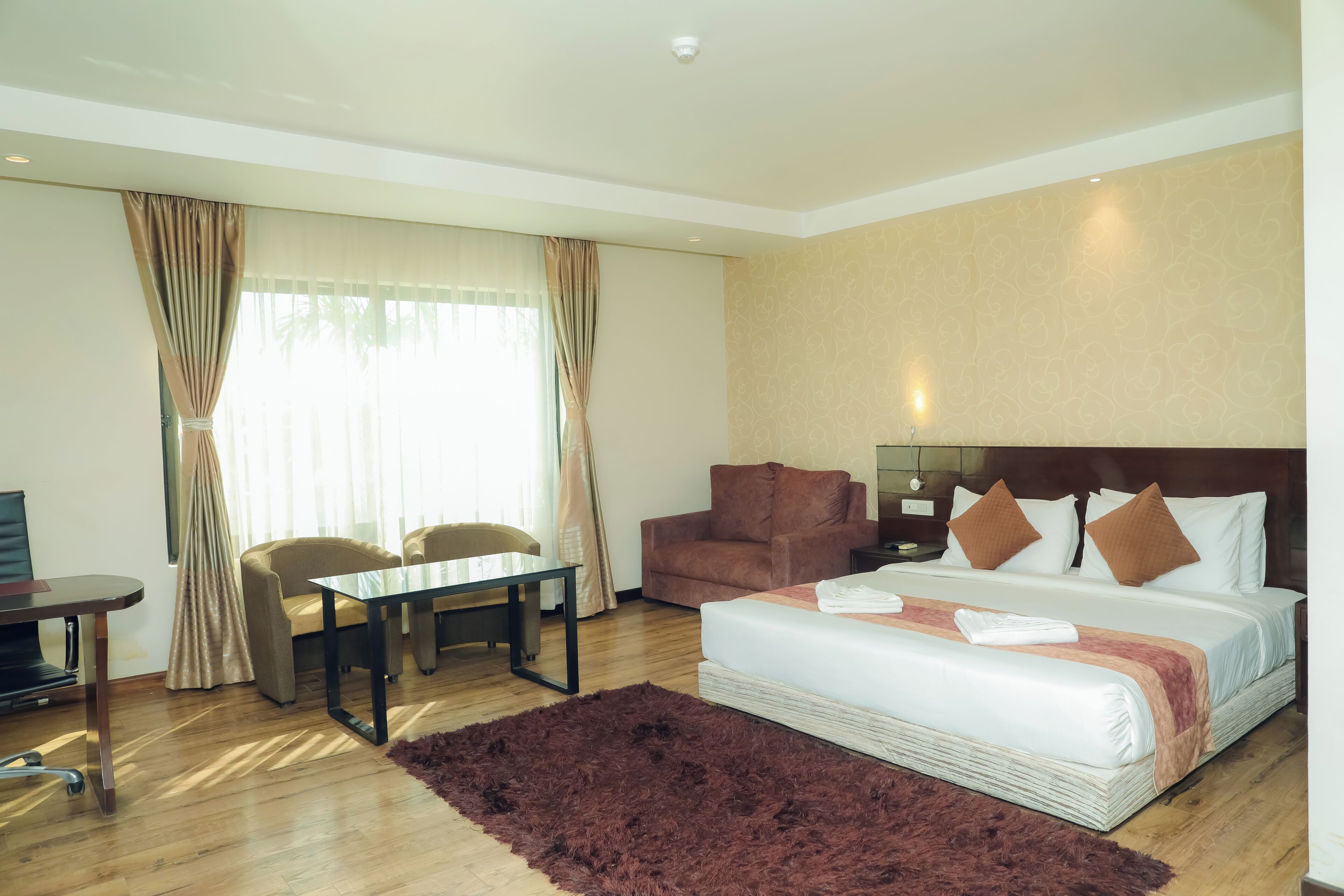 Habitación de hotel con cama, escritorio, dos sillas, televisor y una ventana grande con cortinas.