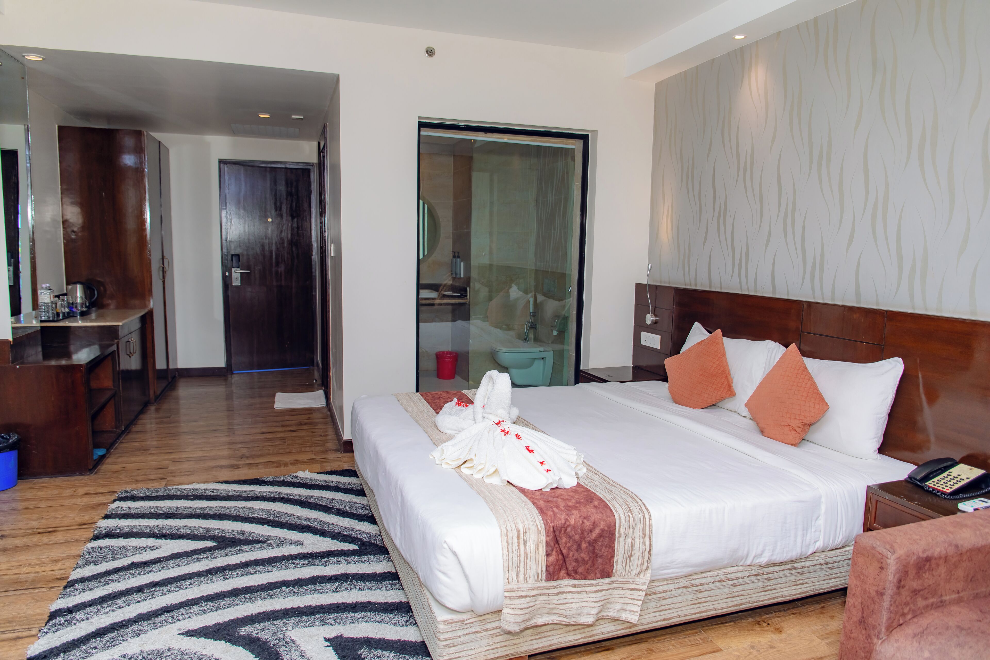 Habitación de hotel con una cama grande, cabecera de madera, mesita de noche con un teléfono y vista al baño a través de una puerta de cristal.