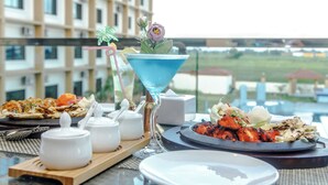 A dining table with a variety of dishes, including grilled meat and vegetables, and a blue cocktail glass with a flower. - Bodhi Redsun – Gateway to Lumbini, Comfort in Bhairahawa (Basantapur)