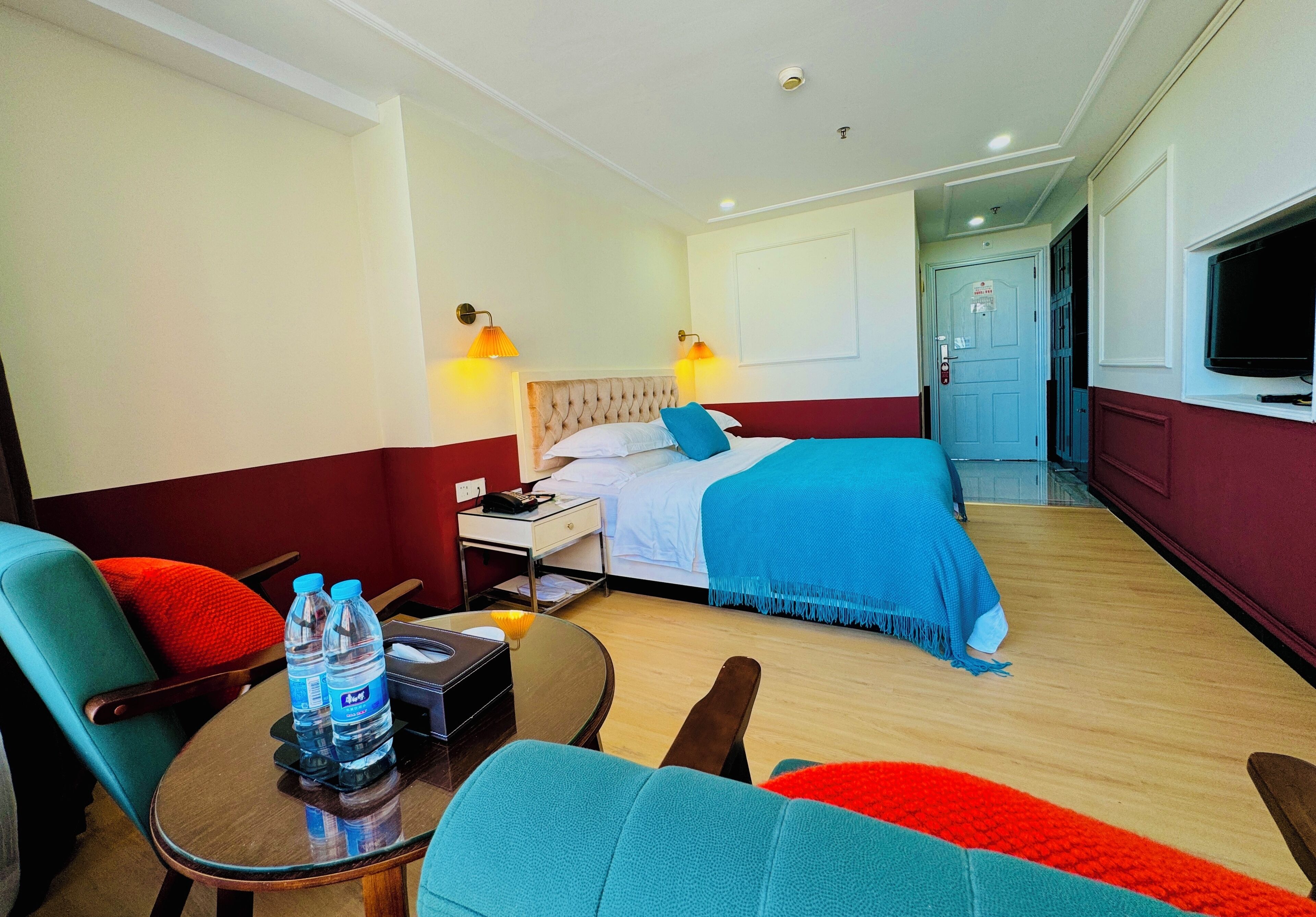 A hotel room with a bed, a desk, a chair, a table with water bottles, and a TV.
