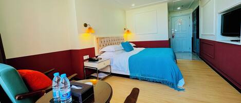 A hotel room with a bed, a desk, a chair, a table with water bottles, and a TV.