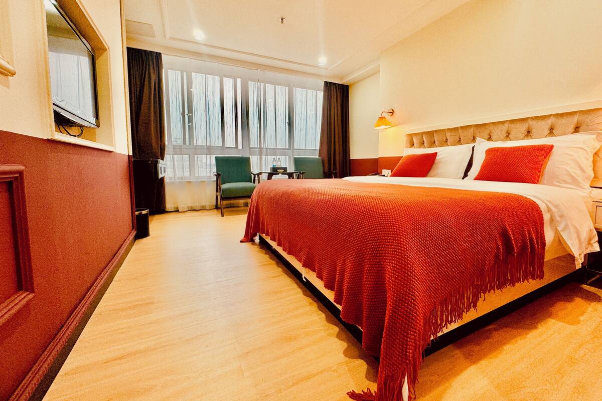 A hotel room with a large bed, a TV mounted on the wall, a window with curtains, and a small table with chairs.