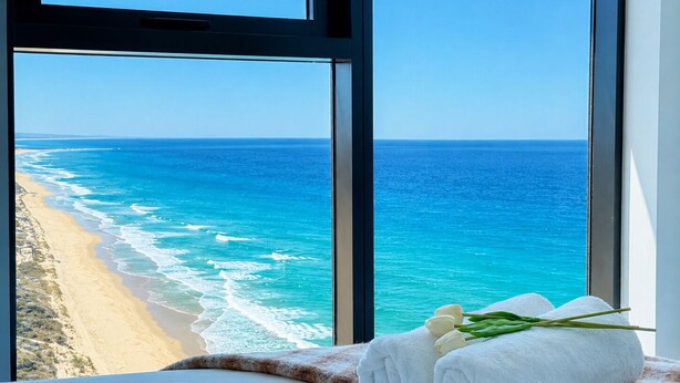 A room with a large window overlooking a beach and ocean, a bed with white linens, and a rolled white towel with green leaves. - Ocean Beachfront Luxe Escape at Surfers Paradise Parking (Surfers Paradise)