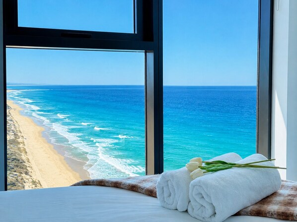 A room with a large window overlooking a beach and ocean, a bed with white linens, and a rolled white towel with green leaves.