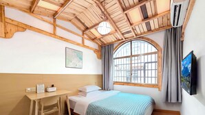 Quarto com teto de madeira, cama com manta estampada, escrivaninha com cadeira, janela com cortinas e televisão fixada na parede.