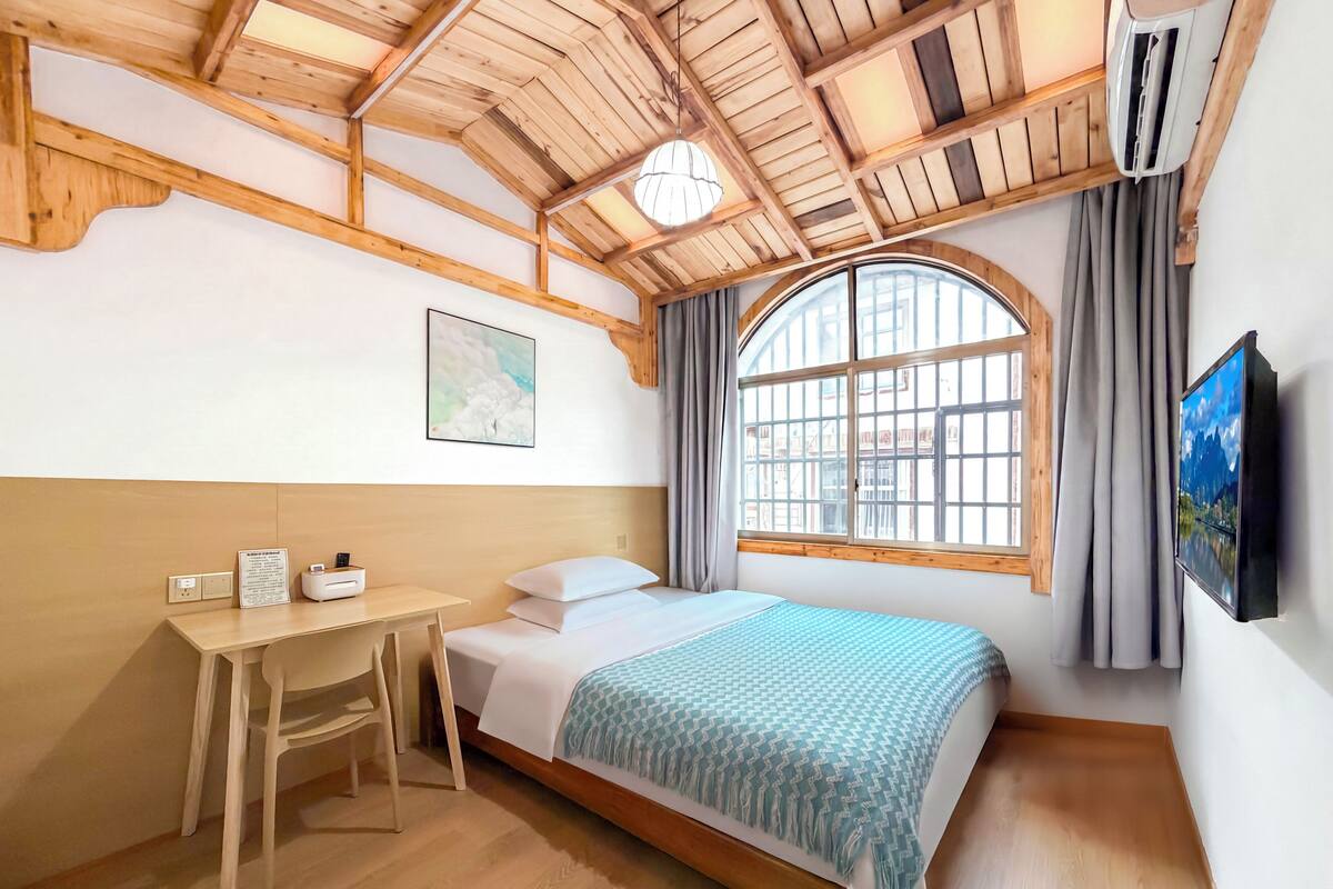 Un dormitorio con techo de madera, una cama con manta estampada, un escritorio con silla, una ventana con cortinas y un televisor montado en la pared.