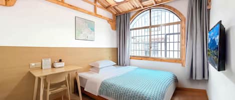 A bedroom with a wooden ceiling, a bed with a patterned blanket, a desk with a chair, a window with curtains, and a wall-mounted TV.