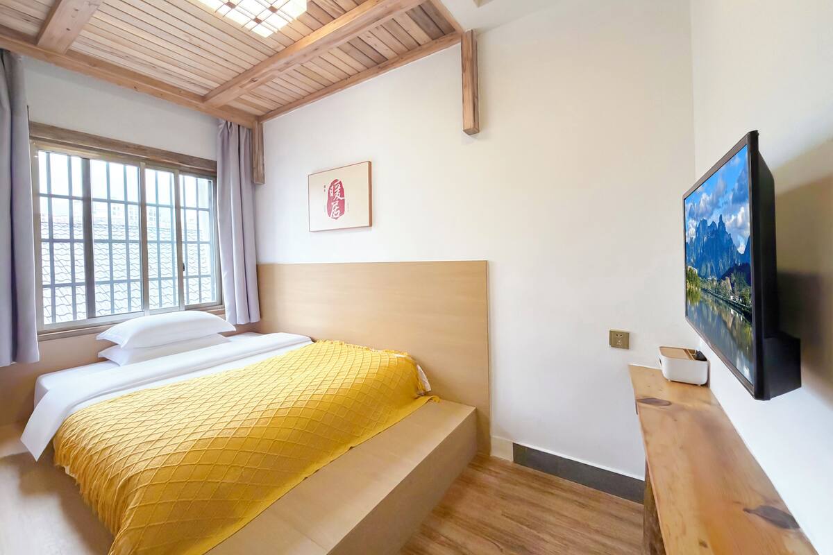 Un dormitorio con cama, cabecera de madera, ventana con cortinas, televisor montado y un cuadro enmarcado en la pared.