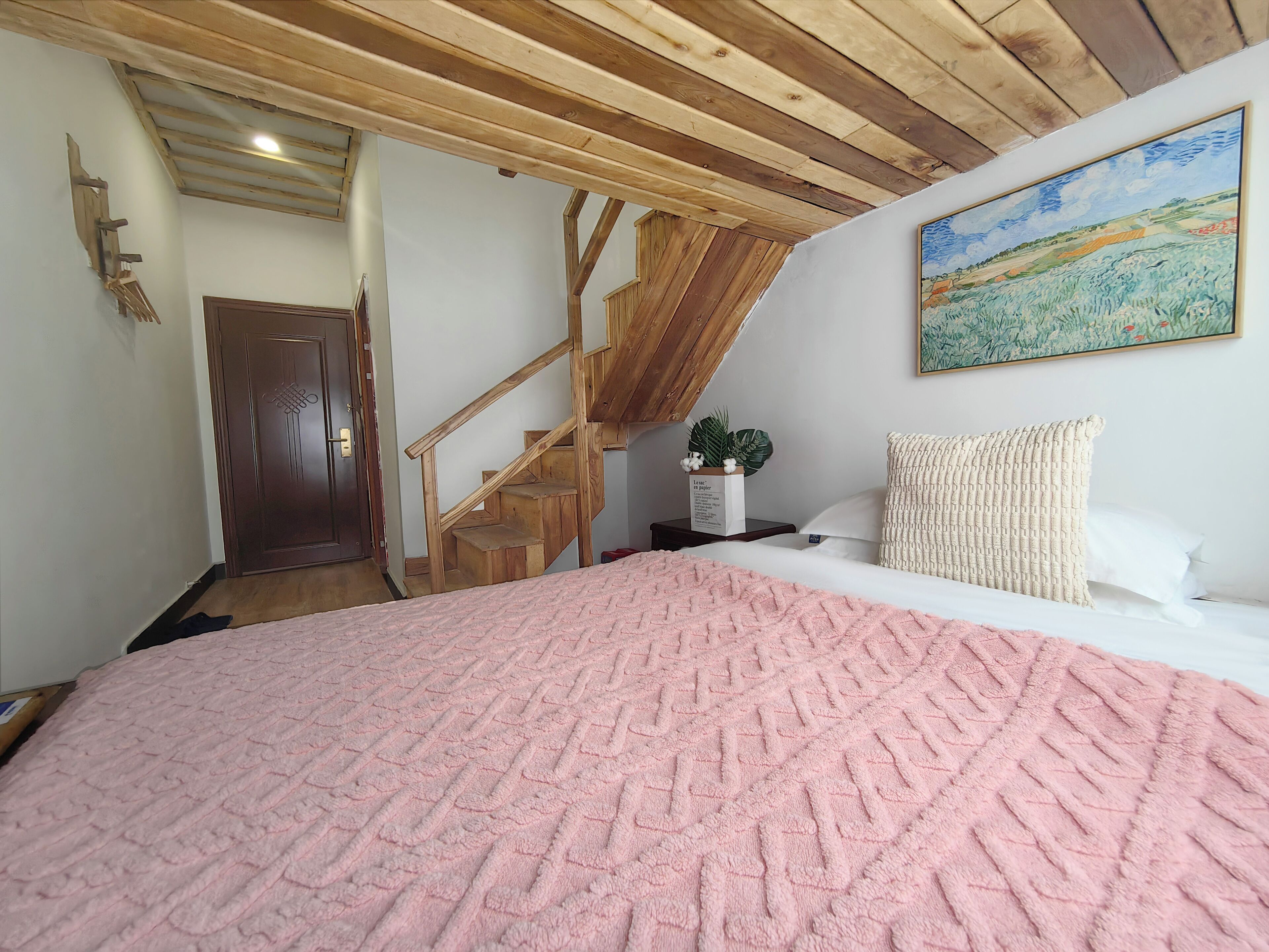 A bedroom with a wooden staircase, a bed with a pink textured blanket, a white pillow, and a painting on the wall.
