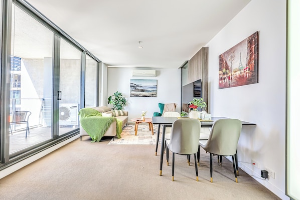 A modern living room with a sofa, a dining table with chairs, a large window, and a painting on the wall.
