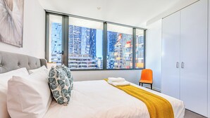 Ein modernes Hotelzimmer mit einem großen Bett, Blick auf die Stadt und einem leuchtend orangefarbenen Sessel.