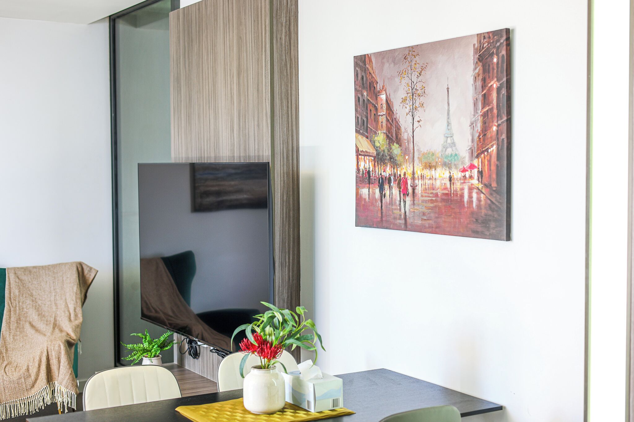 A modern hotel room with a dining area, a large painting of a city street, a flat-screen TV, and a plant on the table.