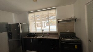 Een kleine keuken met een koelkast, fornuis, gootsteen en raam met uitzicht op een woonwijk.