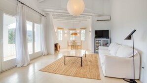 A spacious living room with a white sofa, a coffee table, and a TV. There is a large window with curtains and a view of greenery outside. - CALMA: Spectacular holiday rental villa with private pool and sea views in Sa Riera, Begur. (Begur)