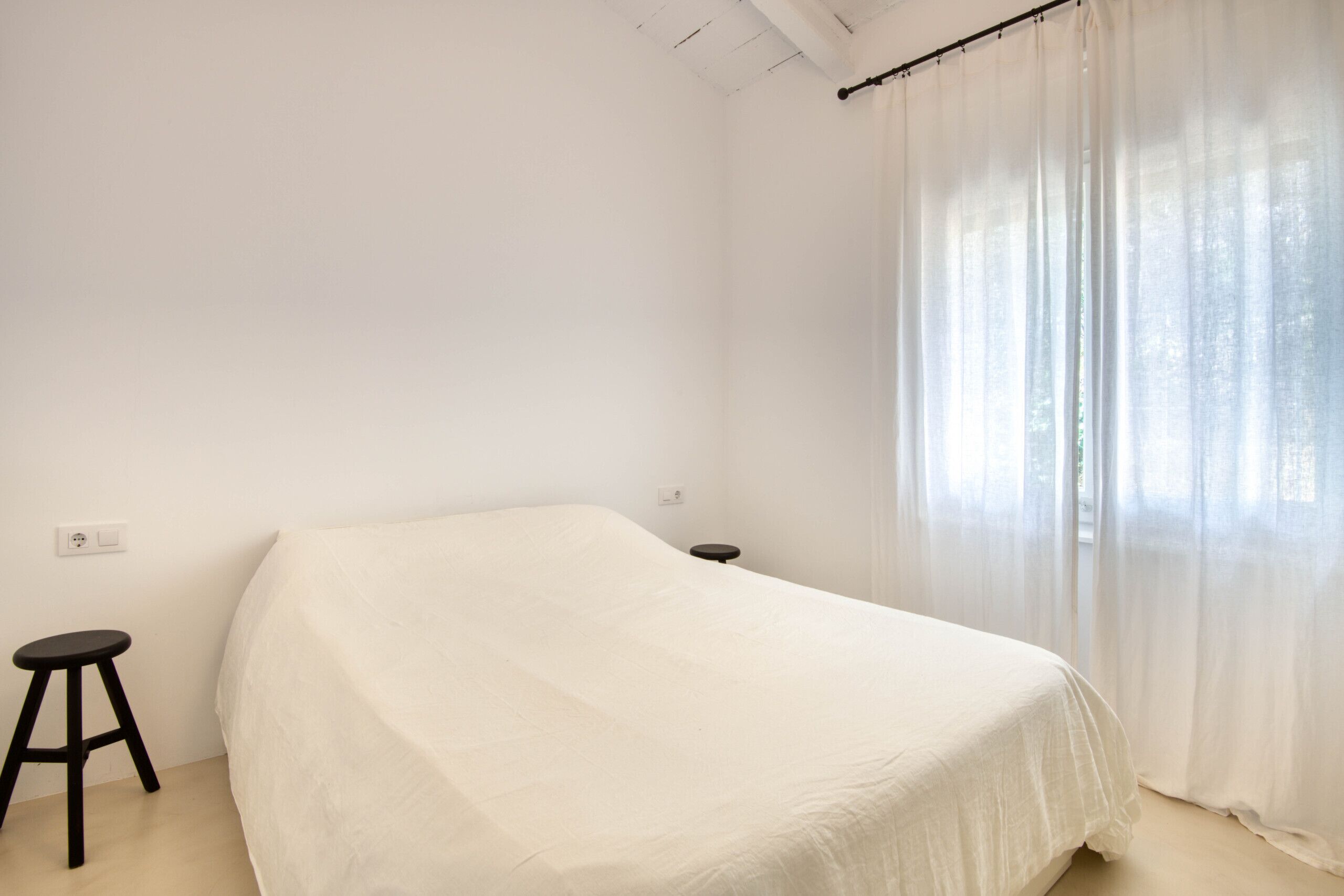 A simple, unadorned room with a bed, a small stool, and a window with sheer curtains.