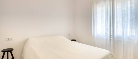 A simple, unadorned room with a bed, a small stool, and a window with sheer curtains.