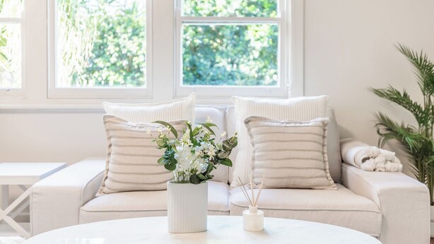 Ett mysigt vardagsrum med en vit soffa, randiga kuddar, en vas med blommor och ett litet bord.