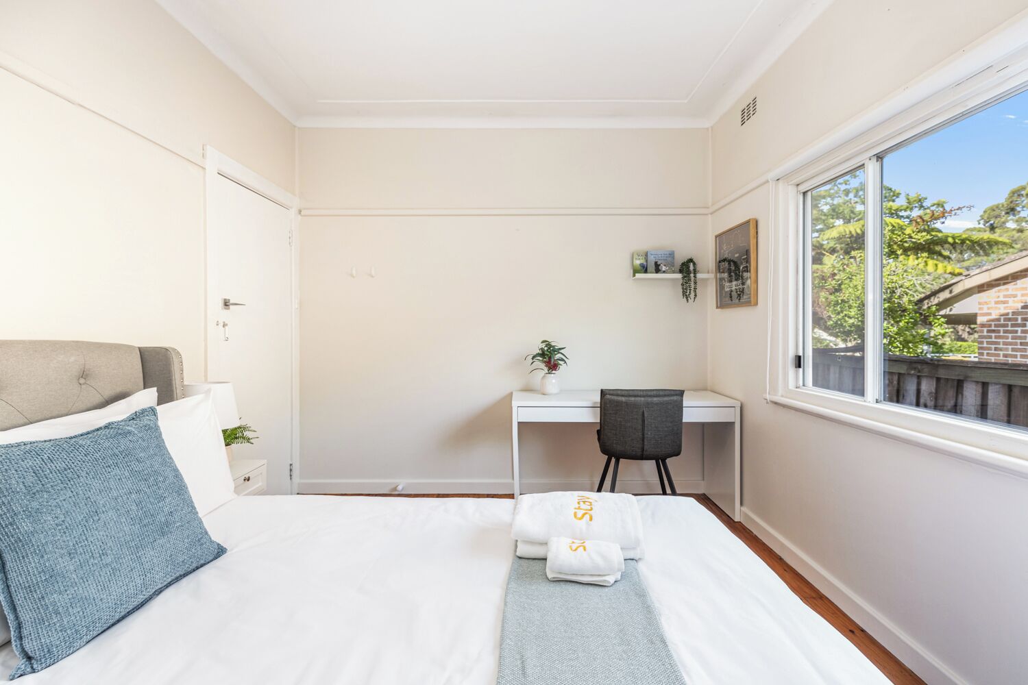 A bedroom with a bed, a desk, a chair, a window, and a small shelf with a plant.