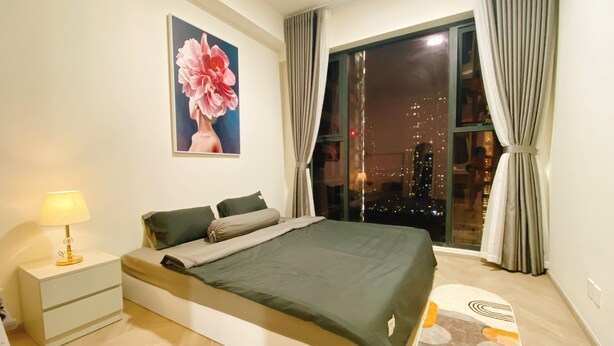 A modern bedroom with a large bed, a nightstand, a lamp, a painting, and a view of the city at night.