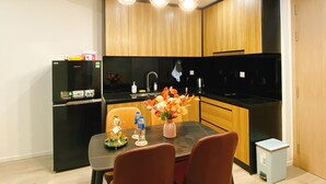 A modern kitchen with wooden cabinets, a black countertop, and a dining table with chairs. There are hanging lights, a refrigerator, and a small trash bin.