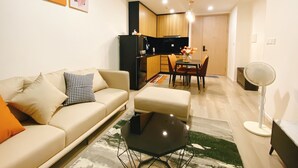 A modern living room with a sofa, a coffee table, and a dining area with chairs and a table. There is a kitchen area with cabinets and a refrigerator.