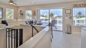 Een moderne woonkamer met een bank, eettafel en stoelen, grote ramen met uitzicht op het water, en een 'BEACH' bord aan de muur.