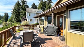 Een houten terras met buitenmeubilair, een overdekte barbecue en een uitzicht op een huis op de achtergrond.