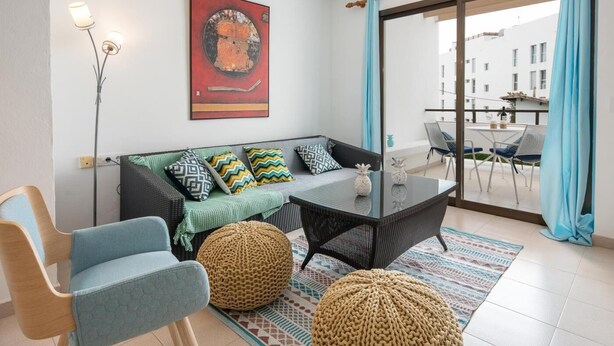 Ein modernes Wohnzimmer mit einer Couch, einem Couchtisch und einem Sessel. An der Wand hängt ein Gemälde und vom Balkon aus hat man einen Tisch und Stühle.