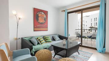 Una sala de estar moderna con un sofá, una mesa de centro y una silla. Hay un cuadro en la pared y un balcón con una mesa y sillas.