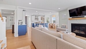 A modern living room with a beige sofa, a TV mounted above a fireplace, and a kitchen with blue cabinets and a central island. - Ideally located, game room, finished basement, fenced yard, lawn games, coffee (Middletown)