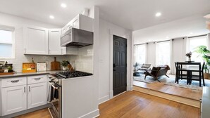 A modern kitchen with white cabinets, a gas stove, and a dark door leading to a living area with a sofa and a dining table. - ★HostWise Stays★ Stylish War Streets Retreat | Easy Walk to Stadiums & Dining (Pittsburgh)
