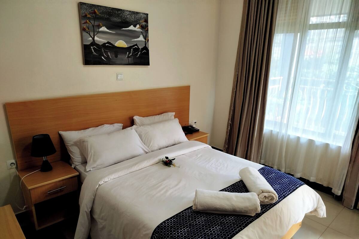 Une chambre d’hôtel avec un grand lit, une table de chevet avec une lampe, un tableau au mur et une fenêtre avec des rideaux.