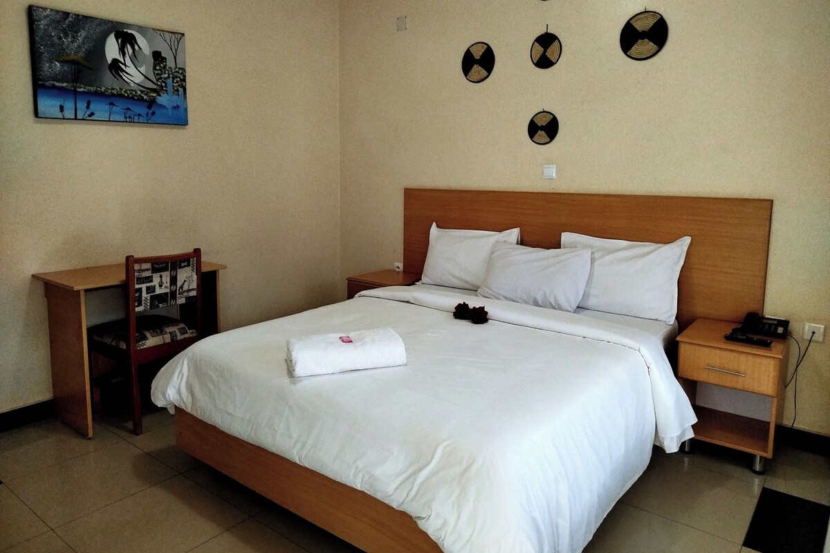A hotel room with a wooden bed, white bedding, a nightstand with a phone, a chair, a small table, and a wall-mounted artwork.