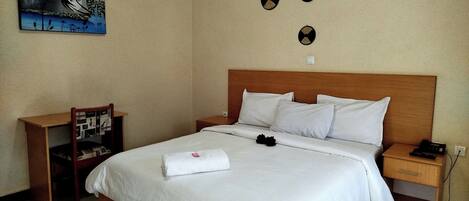 A hotel room with a wooden bed, white bedding, a nightstand with a phone, a chair, a small table, and a wall-mounted artwork.