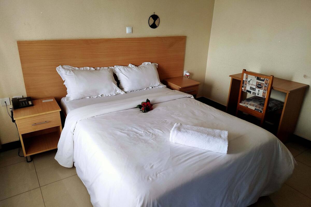 Une chambre d’hôtel avec un grand lit, une tête de lit en bois, une table de chevet avec un téléphone, une petite table de nuit et une chaise avec un coussin à motifs.