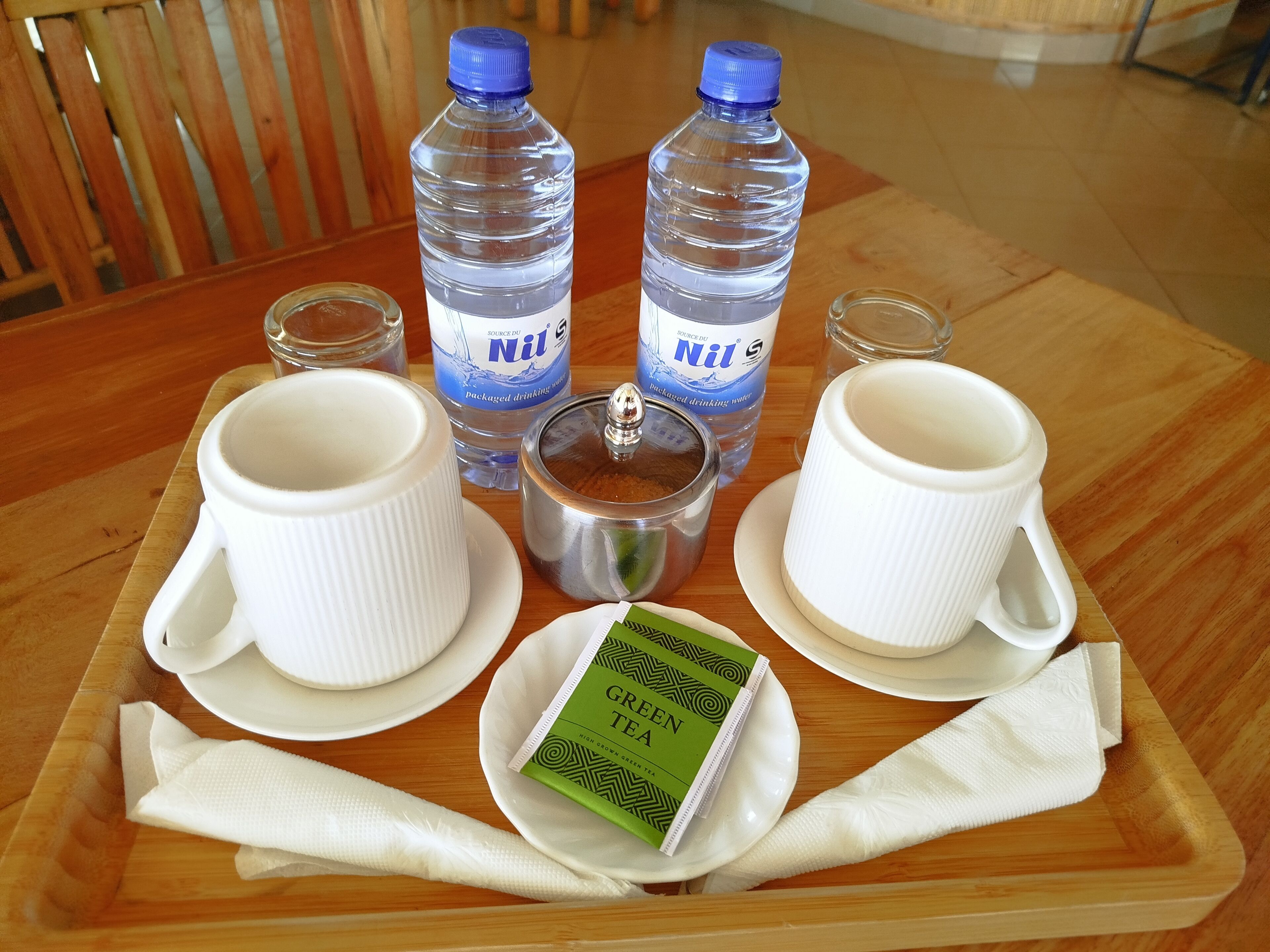 Una bandeja con dos tazas blancas, dos botellas de agua, un recipiente plateado y una bolsita de té verde sobre una mesa de madera.
