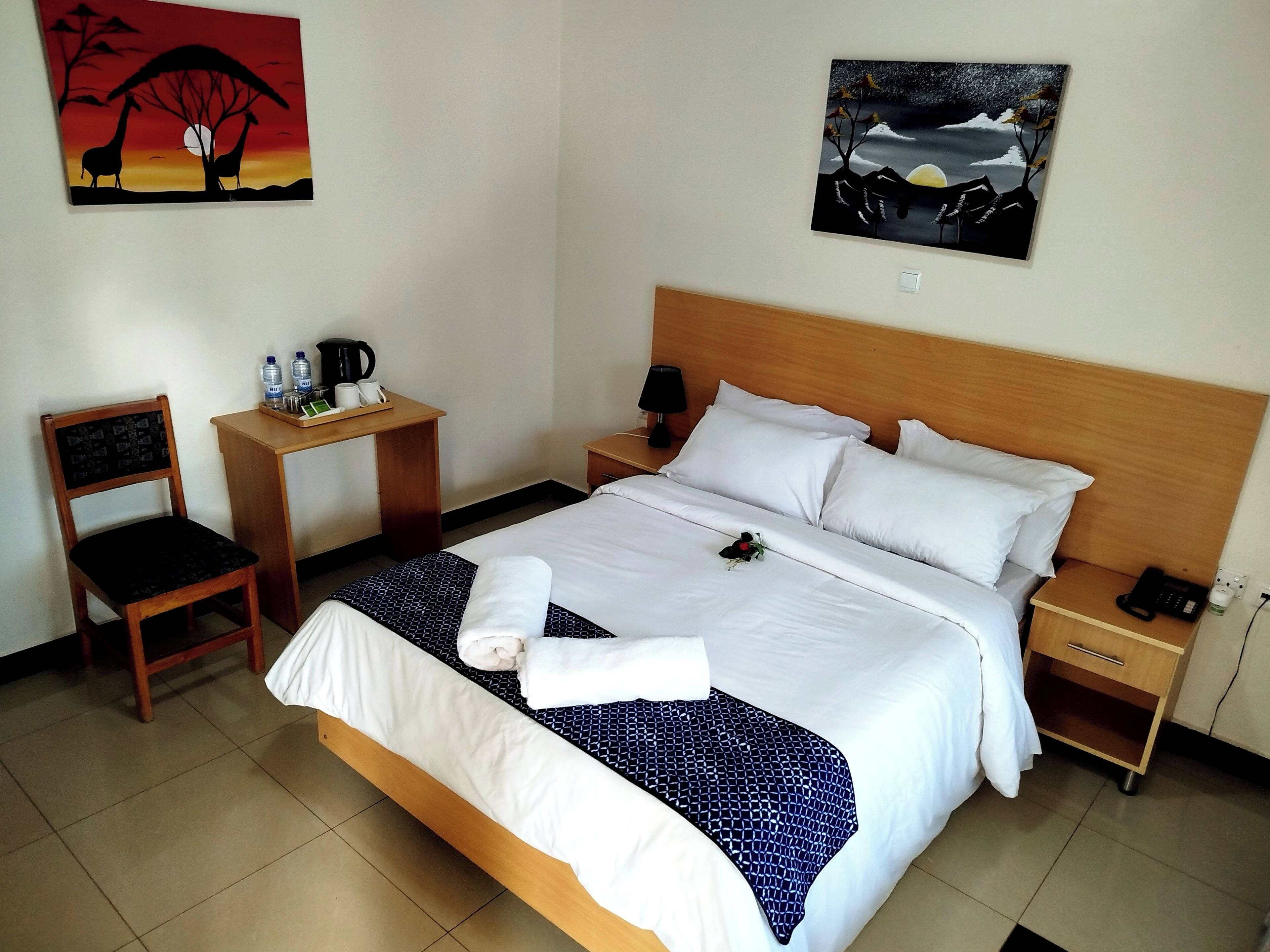 A hotel room with a large bed, a wooden nightstand, a desk with a chair, and two paintings on the walls.