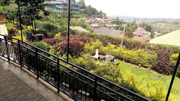 A balcony with a view of a garden, a building with multiple floors, and a distant cityscape.