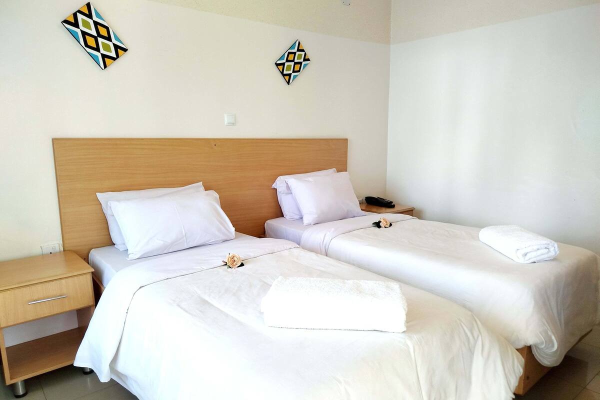 Une chambre d’hôtel avec deux lits, du linge de lit blanc, une tête de lit en bois, une table de chevet, un téléphone et des œuvres d’art décoratives au mur.