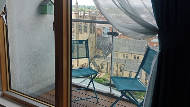 A balcony with two green chairs, a small birdhouse, and a view of historic buildings. - 1 Bed Flat - Sleeps 5 - Pets - Garden - Parking (Manchester)