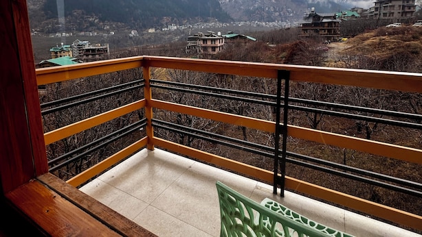 A balcony with a wooden railing overlooking a mountainous landscape with buildings in the distance. - Namgials by Sky Group (Manali)