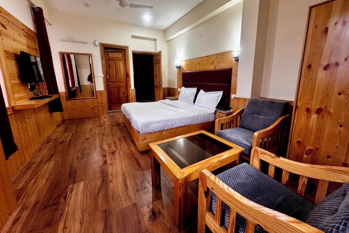 A wooden-floored room with a bed, a chair, a table, and a television on a shelf.