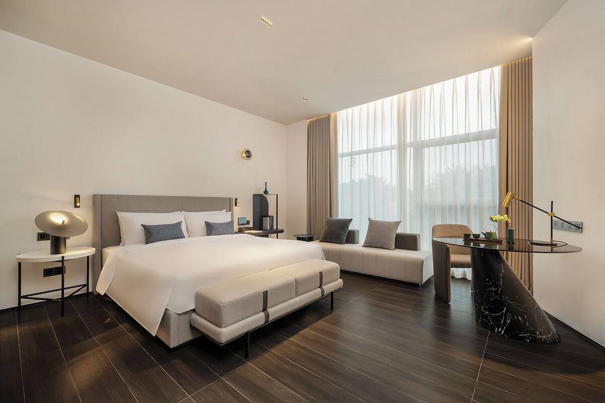 A modern hotel room with a large bed, a sofa, a small table, and a view of the city through large windows.