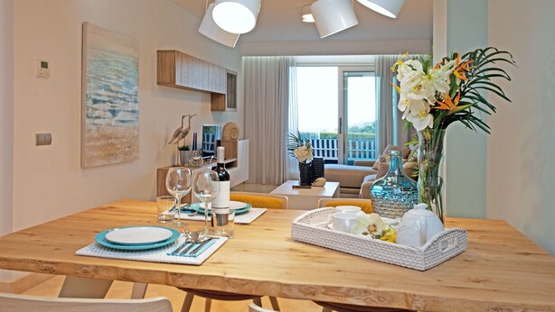 Ein Essbereich mit einem Holztisch, gedeckt für vier Personen, einer Blumenvase und Blick auf das Wohnzimmer und den Balkon.
