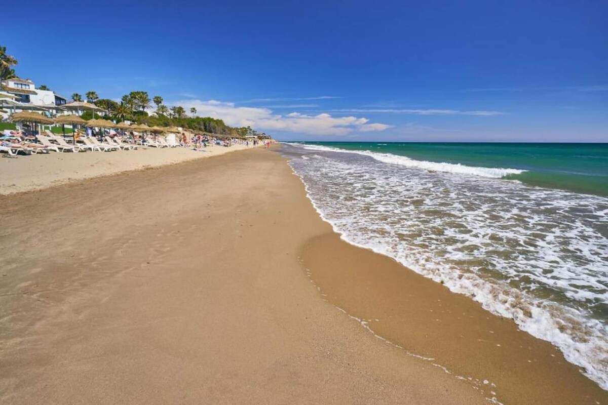 A beach with a clear view of the ocean, a sandy shore, and a row of beachside buildings with umbrellas.