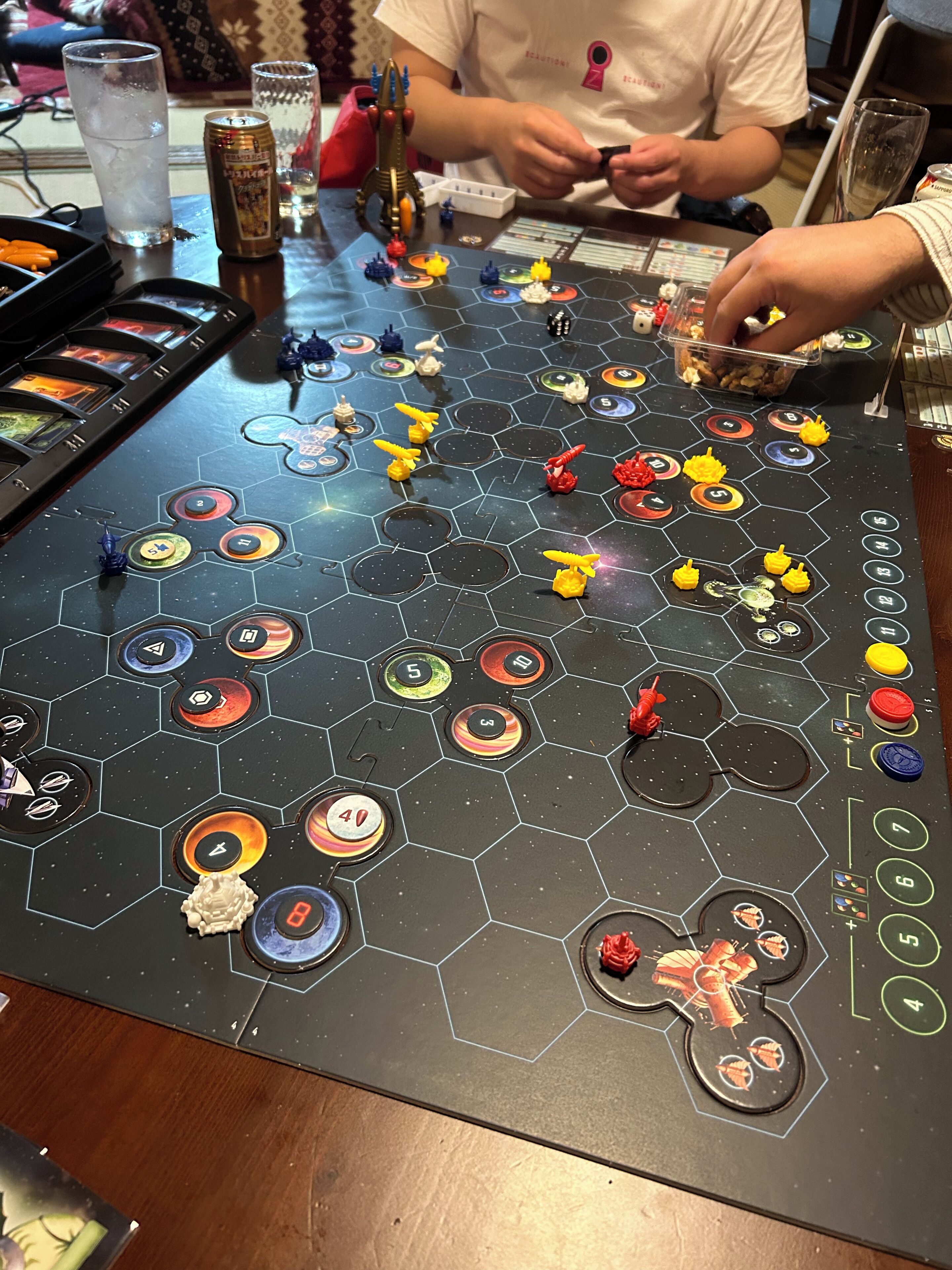 Un juego de mesa con losetas hexagonales y casillas numeradas, una lata de refresco y un objeto con forma de cohete sobre una mesa.