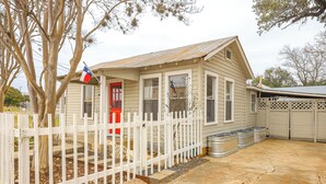 Une petite maison à un étage, avec une porte rouge, une clôture blanche en piquets et un drapeau.