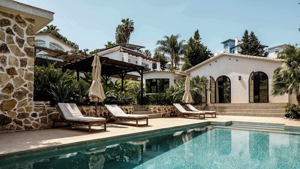 Un área de piscina con sillones de descanso, una pérgola y un edificio con ventanas de arco.