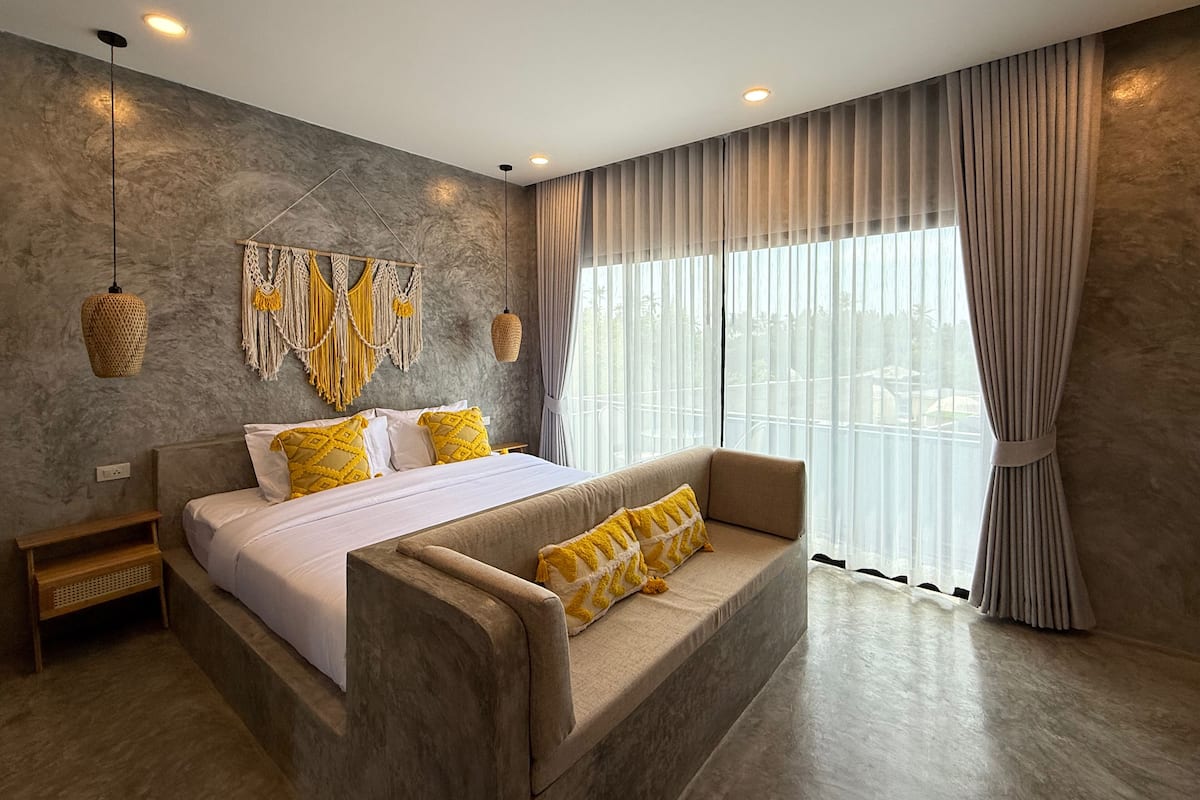 A modern bedroom with a large bed, a sofa, a macrame wall hanging, and a view through sheer curtains.
