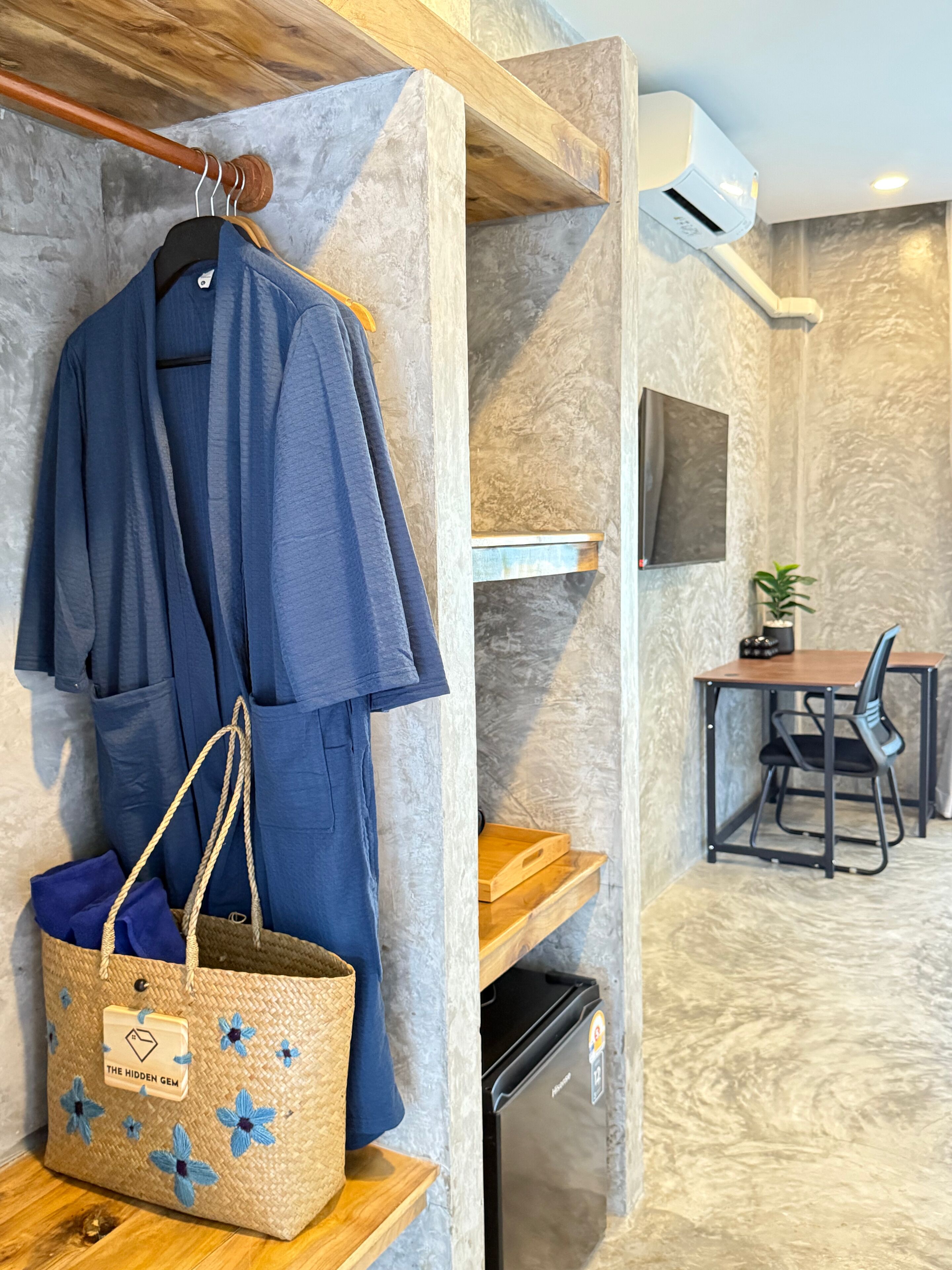 Habitación de hotel con armario de madera, bata colgada, bolso tejido, una mesita con una silla y equipo de aire acondicionado instalado.